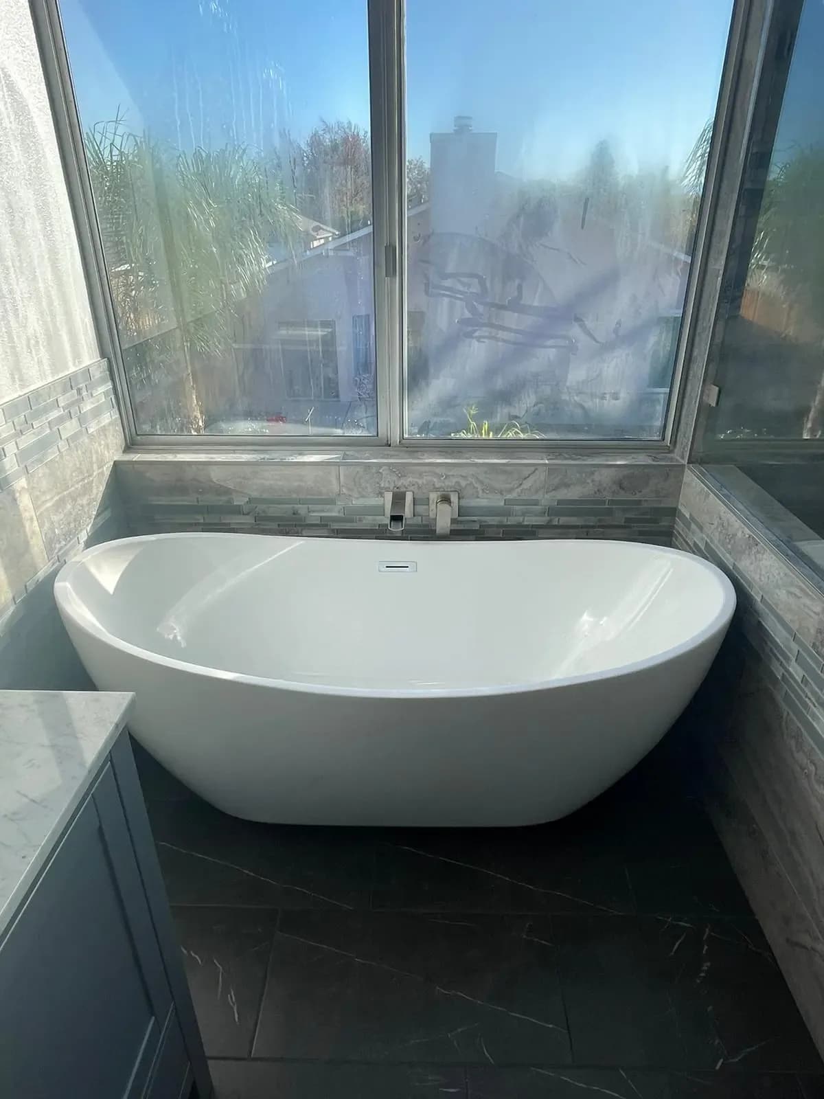 Freestanding tub set under a large window with dark tile surround